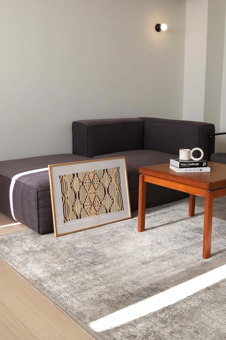 K&ouml;nnte beinhalten: Ein gerahmtes Kunstwerk mit einem geometrischen Muster in Beige-, Burgunder- und Cremet&ouml;nen liegt auf einem grauen Teppich. Das Kunstwerk wird in einem modernen Wohnzimmer pr&auml;sentiert, zusammen mit einem dunkelgrauen Ecksofa und einem Holztisch.