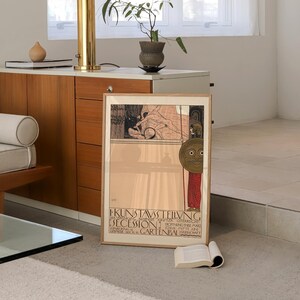 May include: A framed vintage poster with a black and white illustration of a woman holding a spear and a shield with a face on it. The poster is titled "KUNSTAUSTELLUNG DER  SEZESSION" and is for an exhibition of art in Vienna, Austria. The poster is in a wooden frame and is hanging on a wall.