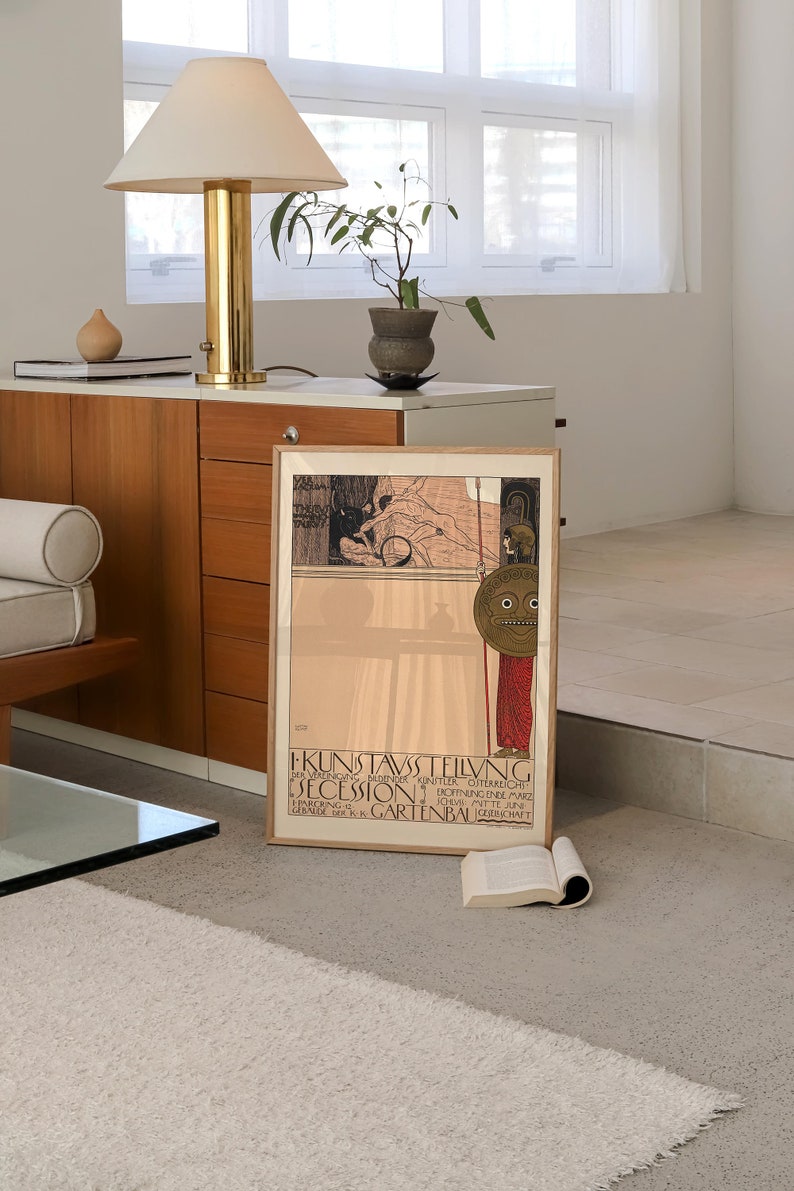 May include: A framed vintage poster with a black and white illustration of a woman holding a spear and a shield with a face on it. The poster is titled "KUNSTAUSTELLUNG DER  SEZESSION" and is for an exhibition of art in Vienna, Austria. The poster is in a wooden frame and is hanging on a wall.