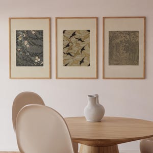 Gallery wall set of three framed Japanese art prints featuring flowering branches, decorative leaf patterns, and a woodland landscape, displayed above a round oak dining table in a warm dining room.