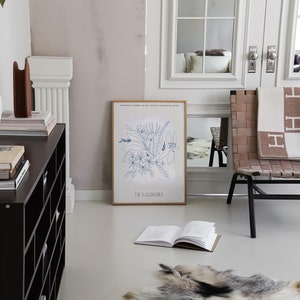 Op de afbeelding: Een ingelijste print van een blauwe lijntekening van bloemen en bladeren met de tekst "DCLASSPARTIE" onderaan. De print hangt aan een witte muur in een kamer met een zwarte plankeneenheid, een witte stoel met een bruine gevlochten zitting en een wit tapijt.