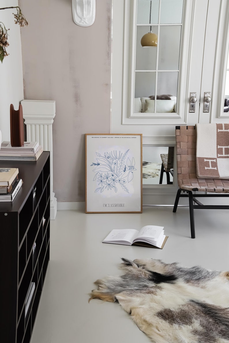 Op de afbeelding: Een ingelijste print van een blauwe lijntekening van bloemen en bladeren met de tekst "DCLASSPARTIE" onderaan. De print hangt aan een witte muur in een kamer met een zwarte plankeneenheid, een witte stoel met een bruine gevlochten zitting en een wit tapijt.