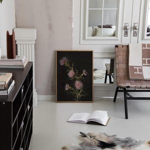 May include: A black and white botanical print of a pink flower with a dark background. The print is framed in a light wood frame and is hanging on a white wall. A black bookshelf with books and a brown object is in the background.