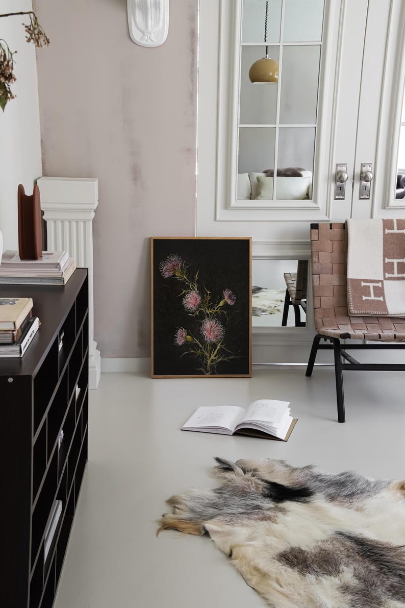 May include: A black and white botanical print of a pink flower with a dark background. The print is framed in a light wood frame and is hanging on a white wall. A black bookshelf with books and a brown object is in the background.