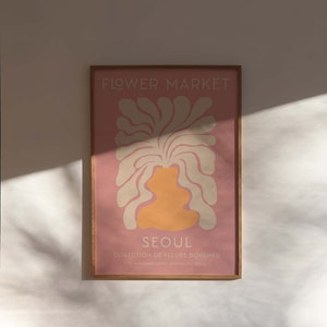 May include: A framed print with a pink background and a stylized illustration of a flower in cream and orange. The text "FLOWER MARKET" is at the top of the print, and the text "SEOUL COLLECTION DE FLEURS BOHEMES 27, GANGNAM-DAERO SEOCHO-GU, SEOUL" is at the bottom.