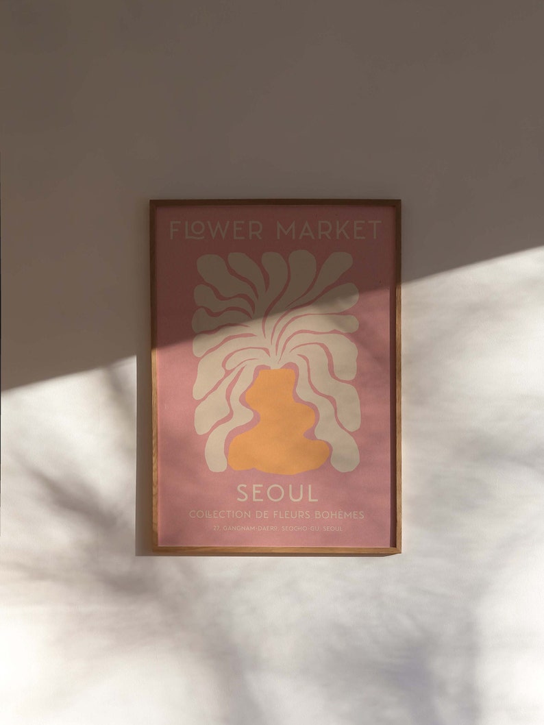 May include: A framed print with a pink background and a stylized illustration of a flower in cream and orange. The text "FLOWER MARKET" is at the top of the print, and the text "SEOUL COLLECTION DE FLEURS BOHEMES 27, GANGNAM-DAERO SEOCHO-GU, SEOUL" is at the bottom.