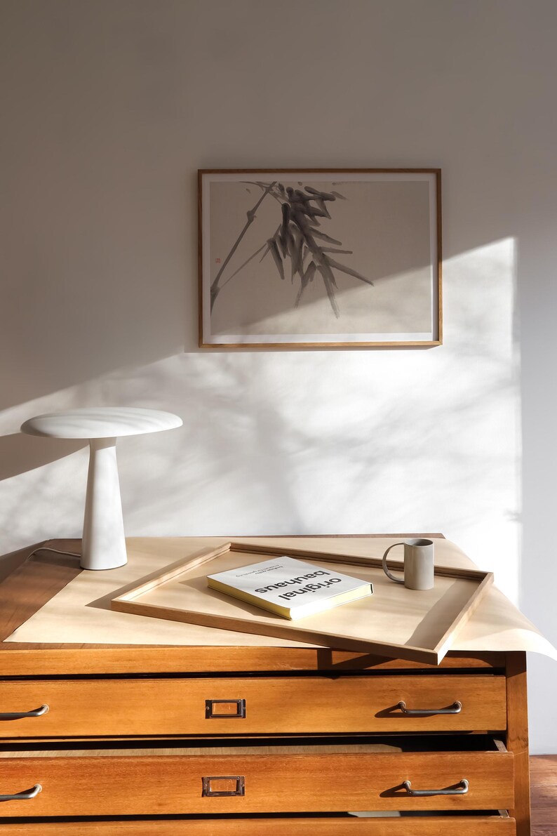 Puede incluir: Escena interior minimalista con una l&aacute;mpara de mesa blanca, una impresi&oacute;n bot&aacute;nica enmarcada y una bandeja de madera con un libro y una taza. La escena est&aacute; ambientada en una c&oacute;moda de madera con cajones, ba&ntilde;ada por la luz natural.