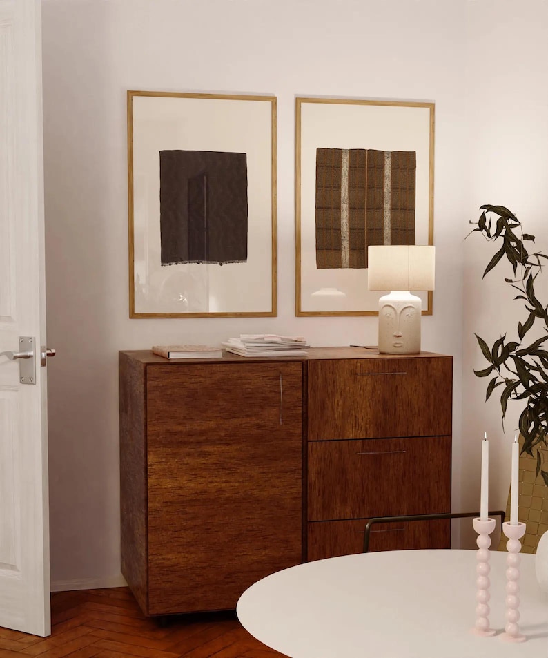 Pu&ograve; includere: Un mobile in legno scuro con un'anta e tre cassetti, sormontato da una lampada bianca con design a volto, due opere d'arte tessile astratte in cornici dorate e due portacandele rosa pallido su un tavolo rotondo bianco.