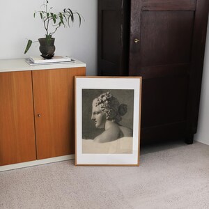 May include: A framed black and white print of a woman's bust sculpture. The sculpture is in profile and the woman has a crown of leaves on her head. The frame is a simple wooden frame.
