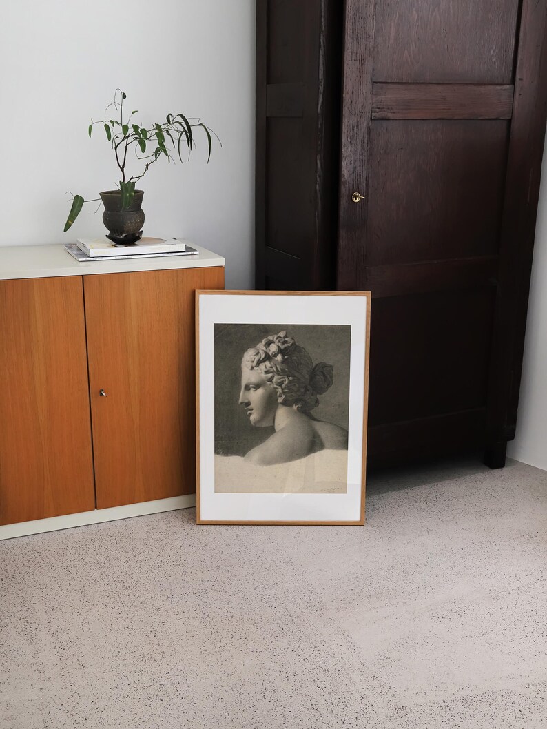 May include: A framed black and white print of a woman's bust sculpture. The sculpture is in profile and the woman has a crown of leaves on her head. The frame is a simple wooden frame.