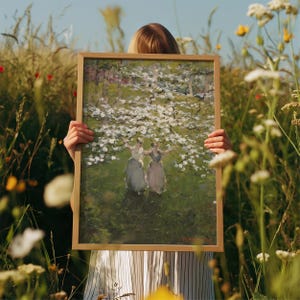 Op de afbeelding: Een ingelijst schilderij van twee vrouwen in witte jurken die onder een bloeiende boom staan. Het schilderij is tegen een groene achtergrond met witte bloemen.