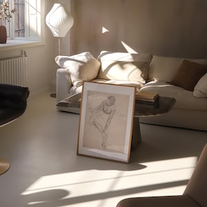 May include: A framed pencil sketch of a dancer in a light-filled living room. The artwork is propped against a small, rustic table in front of a white sofa with beige pillows. A modern white lamp and a beige chair are also visible.