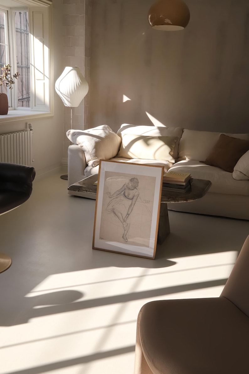 May include: A framed pencil sketch of a dancer in a light-filled living room. The artwork is propped against a small, rustic table in front of a white sofa with beige pillows. A modern white lamp and a beige chair are also visible.