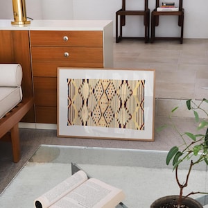K&ouml;nnte beinhalten: Ein gerahmtes Kunstwerk mit einem geometrischen Muster in Beige-, Rot- und Blaut&ouml;nen. Das Kunstwerk lehnt an einer wei&szlig;en Wand. Ein offenes Buch liegt auf einem Glastisch, mit einer kleinen Topfpflanze in der N&auml;he.