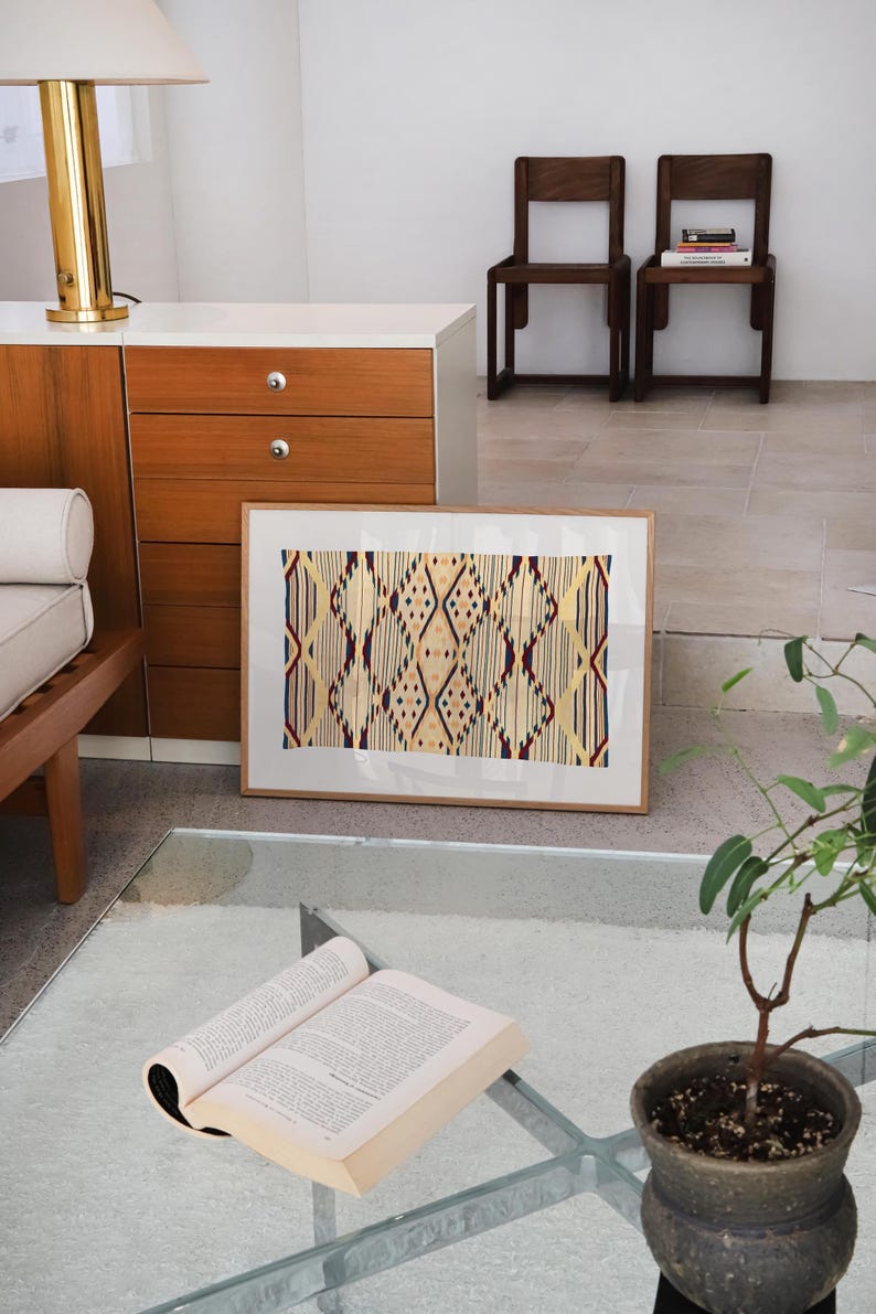 K&ouml;nnte beinhalten: Ein gerahmtes Kunstwerk mit einem geometrischen Muster in Beige-, Rot- und Blaut&ouml;nen. Das Kunstwerk lehnt an einer wei&szlig;en Wand. Ein offenes Buch liegt auf einem Glastisch, mit einer kleinen Topfpflanze in der N&auml;he.