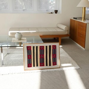 May include: A framed textile art piece with a geometric pattern. The design features vertical stripes in black, tan, and red with blue accents. The artwork is displayed on a white rug in a well-lit room with a modern aesthetic.