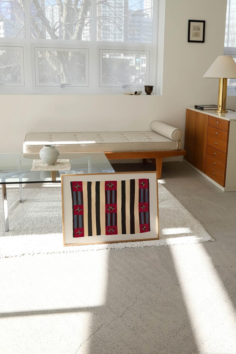 May include: A framed textile art piece with a geometric pattern. The design features vertical stripes in black, tan, and red with blue accents. The artwork is displayed on a white rug in a well-lit room with a modern aesthetic.