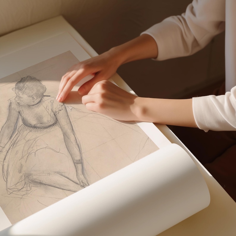 May include: A pencil sketch of a seated ballerina, framed in white, with a rolled white paper in the foreground. The sketch is in shades of gray and depicts a dancer in a flowing dress. The image is lit by natural light.