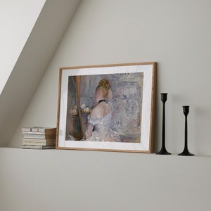 May include: A framed painting of a woman in a white dress, sitting in front of a mirror and fixing her hair. The painting is on a white shelf with two black candle holders.
