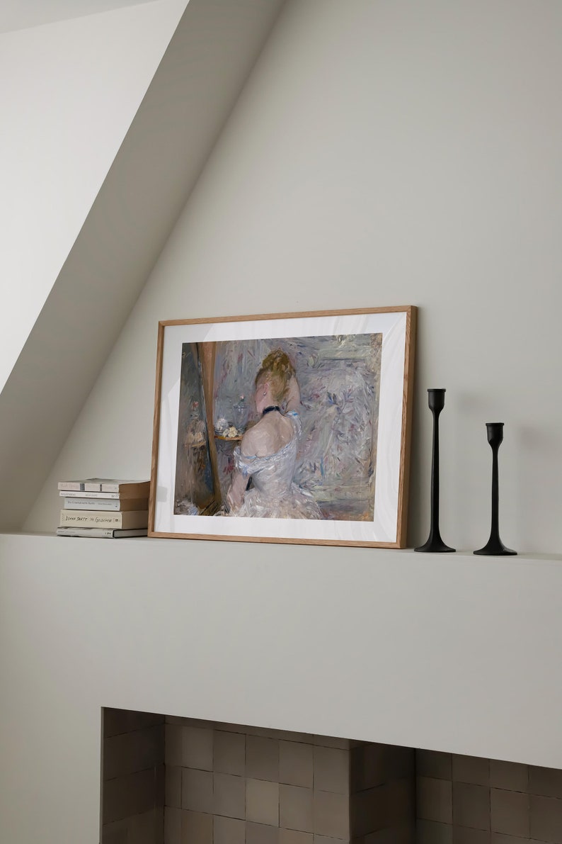 May include: A framed painting of a woman in a white dress, sitting in front of a mirror and fixing her hair. The painting is on a white shelf with two black candle holders.