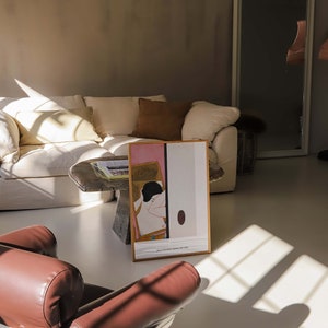 May include: A white sofa with brown cushions in a living room featuring a brown leather armchair and a framed print of a woman in a pink and white dress. The print is on a table with a book on top. The room has a large window with sunlight streaming in.