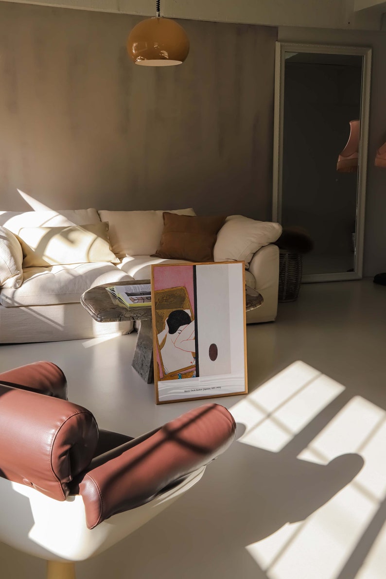 May include: A white sofa with brown cushions in a living room featuring a brown leather armchair and a framed print of a woman in a pink and white dress. The print is on a table with a book on top. The room has a large window with sunlight streaming in.