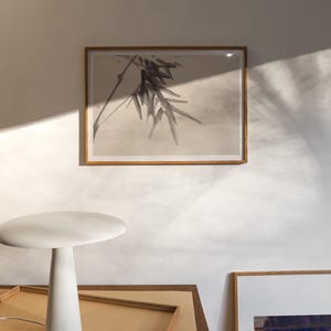 Puede incluir: Interior minimalista con una l&aacute;mpara de mesa blanca y una obra de arte enmarcada. La l&aacute;mpara tiene una base c&oacute;nica y una parte superior redonda. La obra de arte representa una planta. La escena est&aacute; iluminada por luz natural, creando sombras suaves.