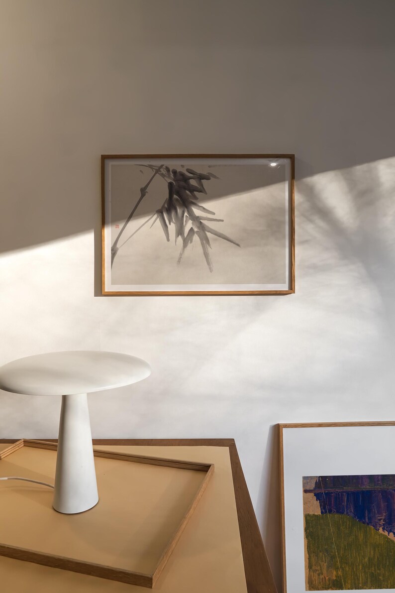 Puede incluir: Interior minimalista con una l&aacute;mpara de mesa blanca y una obra de arte enmarcada. La l&aacute;mpara tiene una base c&oacute;nica y una parte superior redonda. La obra de arte representa una planta. La escena est&aacute; iluminada por luz natural, creando sombras suaves.