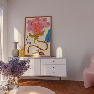 May include: A white dresser with three drawers and gold handles. A framed poster with a colorful abstract design and the text "ZHANG HUOXI" is on the dresser. A white lamp with a curved arch shape is on the dresser. A vase with purple flowers is on a pink round table in front of the dresser.