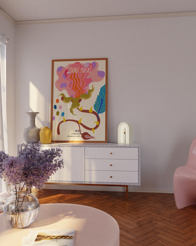 May include: A white dresser with three drawers and gold handles. A framed poster with a colorful abstract design and the text "ZHANG HUOXI" is on the dresser. A white lamp with a curved arch shape is on the dresser. A vase with purple flowers is on a pink round table in front of the dresser.