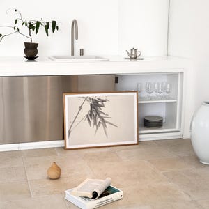 Puede incluir: Una escena de cocina minimalista con una impresi&oacute;n bot&aacute;nica enmarcada, un peque&ntilde;o objeto en forma de pera y un libro abierto. La habitaci&oacute;n tiene una encimera blanca con un fregadero y un armario de acero inoxidable. Tambi&eacute;n se ven una peque&ntilde;a planta y una tetera.