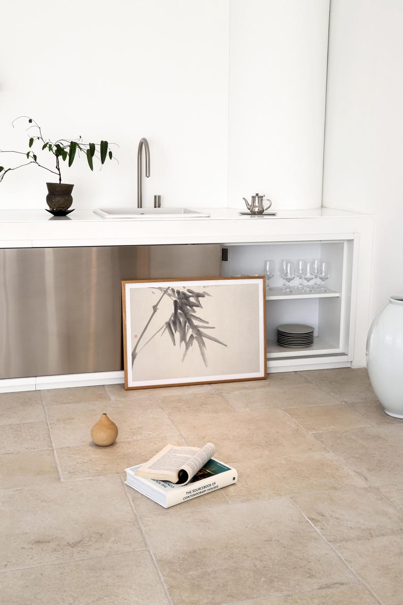 Puede incluir: Una escena de cocina minimalista con una impresi&oacute;n bot&aacute;nica enmarcada, un peque&ntilde;o objeto en forma de pera y un libro abierto. La habitaci&oacute;n tiene una encimera blanca con un fregadero y un armario de acero inoxidable. Tambi&eacute;n se ven una peque&ntilde;a planta y una tetera.
