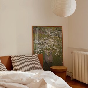 Op de afbeelding: Een wit bed met een wit-zwart gestreept kussen en een wit dekbed. Aan de muur boven het bed hangt een schilderij van een bloeiende boom met drie figuren die ervoor staan. Een houten kruk staat naast het bed.