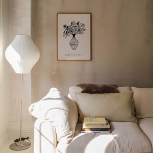 May include: A white couch with a brown throw pillow and a stack of books on the armrest. The couch is in a room with a white lamp and a framed print of a vase of flowers with the text "SUNDAY GALERIE" below the image.