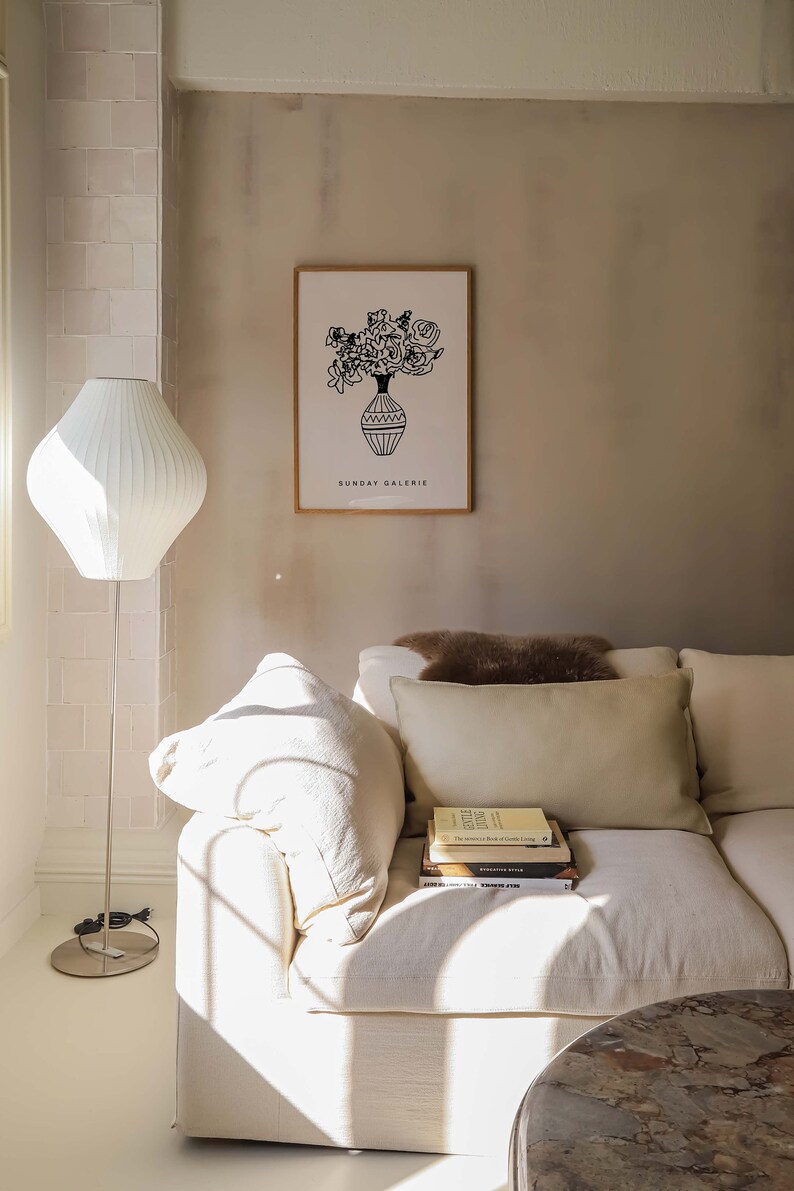 May include: A white couch with a brown throw pillow and a stack of books on the armrest. The couch is in a room with a white lamp and a framed print of a vase of flowers with the text "SUNDAY GALERIE" below the image.