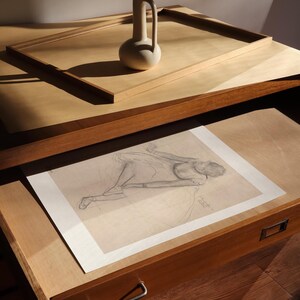 May include: A still life composition featuring a beige ceramic vase with a handle, resting on a wooden surface. Below, a pencil sketch of a figure lies in an open drawer. The scene is bathed in warm light.