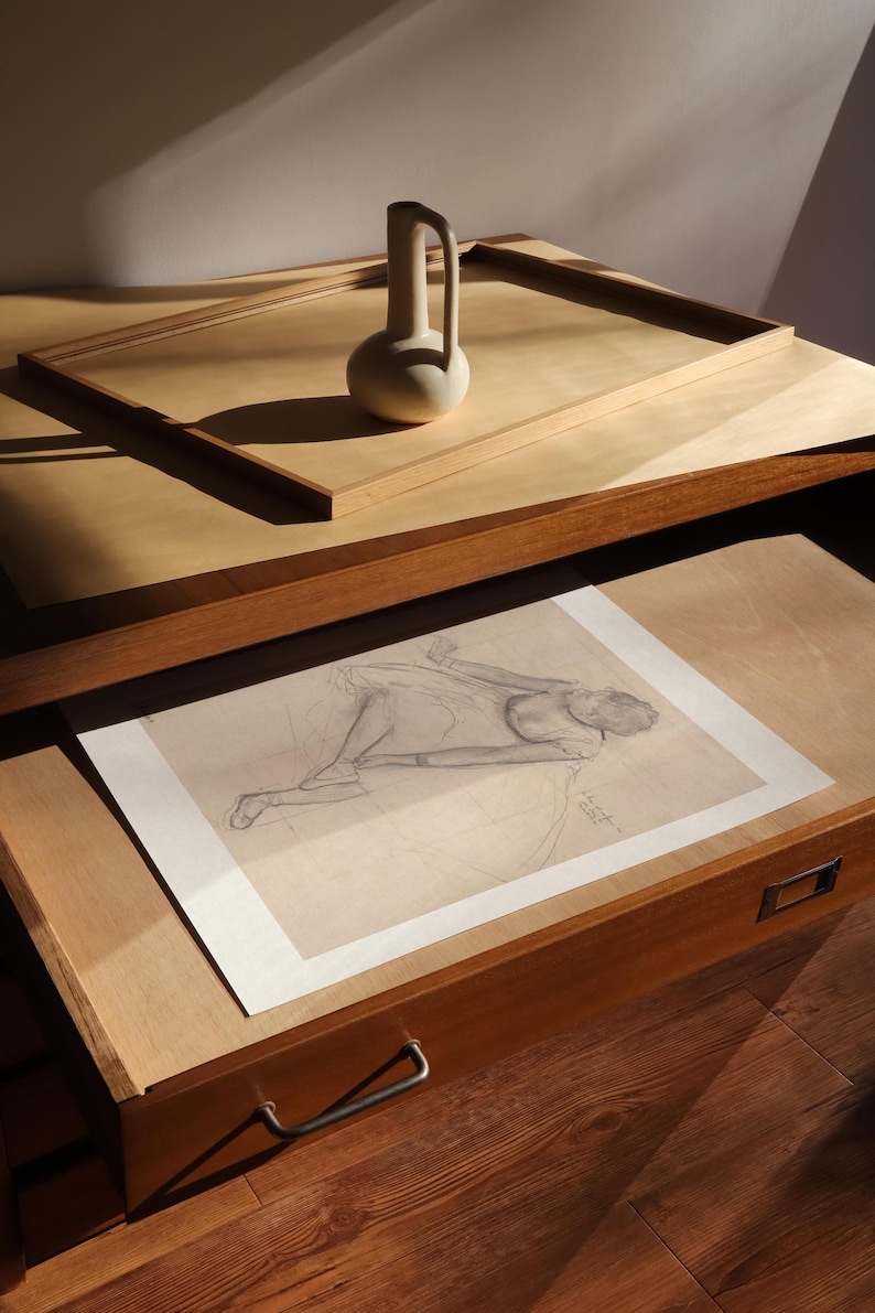 May include: A still life composition featuring a beige ceramic vase with a handle, resting on a wooden surface. Below, a pencil sketch of a figure lies in an open drawer. The scene is bathed in warm light.
