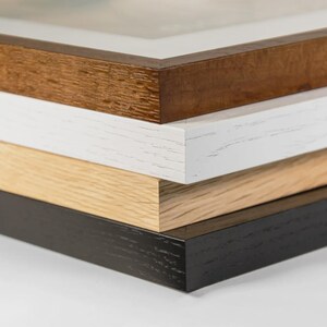 May include: A stack of four picture frames with different wood finishes. The top frame is a dark brown, followed by white, light wood, and black. The frames are stacked, showing the edges and corners.