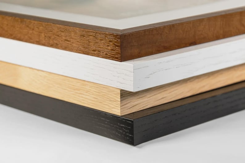May include: A stack of four picture frames with different wood finishes. The top frame is a dark brown, followed by white, light wood, and black. The frames are stacked, showing the edges and corners.