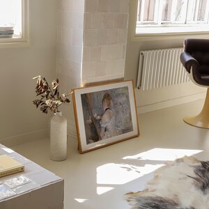 May include: A framed painting of a woman in a white dress, standing in front of a mirror, with a vase of flowers on a table. The painting is hanging on a white wall in a room with a white tiled floor.
