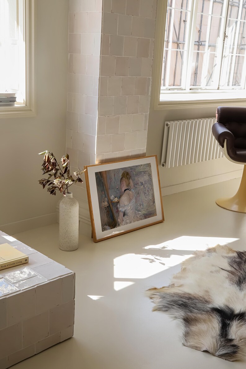 May include: A framed painting of a woman in a white dress, standing in front of a mirror, with a vase of flowers on a table. The painting is hanging on a white wall in a room with a white tiled floor.