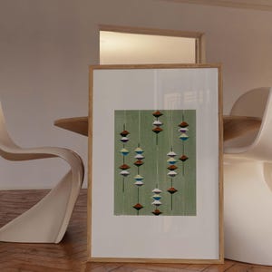 May include: A framed abstract print featuring a green background with vertical lines and colorful, geometric shapes. The artwork is displayed in a light wood frame, leaning against a wall. The print's design includes red, white, blue, and brown elements.