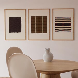 Pu&ograve; includere: Tre stampe artistiche rettangolari incorniciate con motivi tessili astratti in marrone scuro, beige e nero con strisce orizzontali, esposte sopra un tavolo da pranzo rotondo in legno con un vaso in ceramica bianco e due sedie curve.