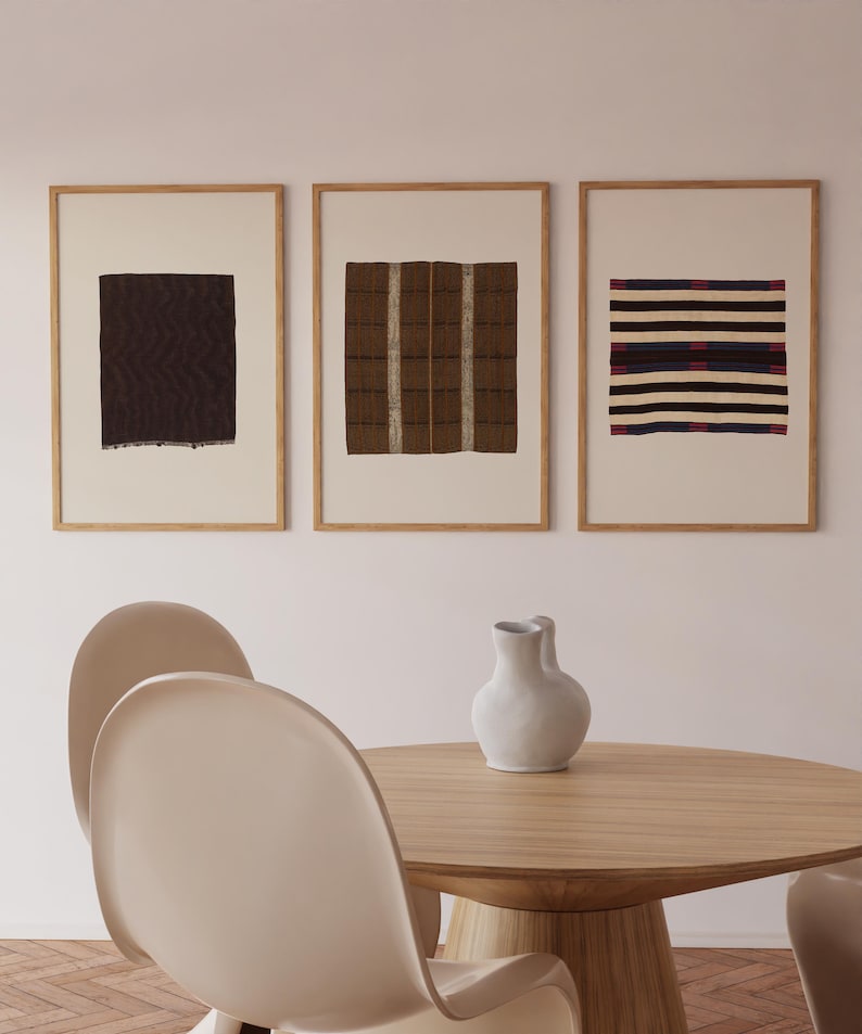 Pu&ograve; includere: Tre stampe artistiche rettangolari incorniciate con motivi tessili astratti in marrone scuro, beige e nero con strisce orizzontali, esposte sopra un tavolo da pranzo rotondo in legno con un vaso in ceramica bianco e due sedie curve.
