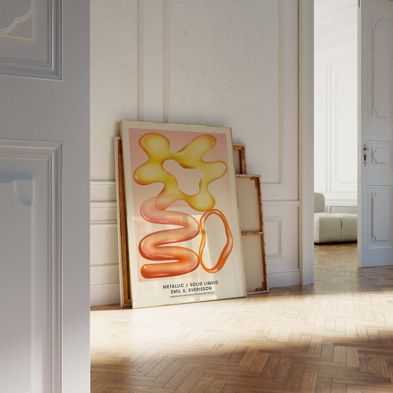 Puede incluir: Una impresi&oacute;n art&iacute;stica enmarcada con formas abstractas en tonos amarillos, naranjas y rosas. La obra de arte, titulada "Metallic / Solid Liquid" de Emil U. Sverisson, se muestra sobre un fondo rosa claro. La impresi&oacute;n est&aacute; apoyada contra una pared en una habitaci&oacute;n con suelo de madera.