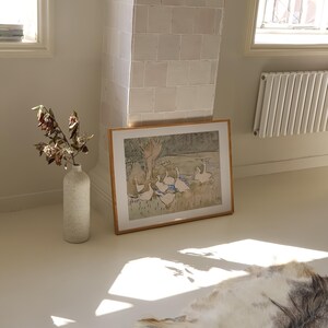 May include: A framed horizontal print featuring a watercolor illustration of a group of white geese in a grassy field with trees, placed next to a tall, speckled, off-white ceramic vase containing dried branches on a smooth, light-colored floor.