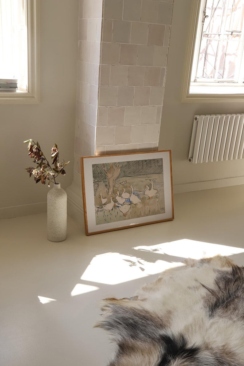 May include: A framed horizontal print featuring a watercolor illustration of a group of white geese in a grassy field with trees, placed next to a tall, speckled, off-white ceramic vase containing dried branches on a smooth, light-colored floor.