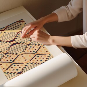 K&ouml;nnte beinhalten: Nahaufnahme eines gemusterten Textils mit beigem Hintergrund und geometrischen Designs in Marineblau, Burgunderrot und Pfirsich. Das Textil wird auf einer wei&szlig;en Oberfl&auml;che bearbeitet. Das Design zeigt Linien, Rauten und andere Formen.
