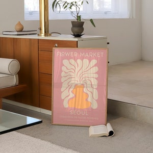 May include: A pink poster with a white and orange abstract floral design. The poster has the text "FLOWER MARKET" and "SEOUL" in white. The text below "SEOUL" reads "COLLECTION DE FLEURS BOHEMES" and "27, BOULEVARD SEOUL, 00000 SEOUL".