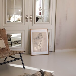 May include: A framed pencil sketch of a seated figure, leaning against a wall. The artwork is displayed on a light-colored floor, with a natural fur rug in the foreground. A woven leather chair and a set of double doors with glass panels are also visible.